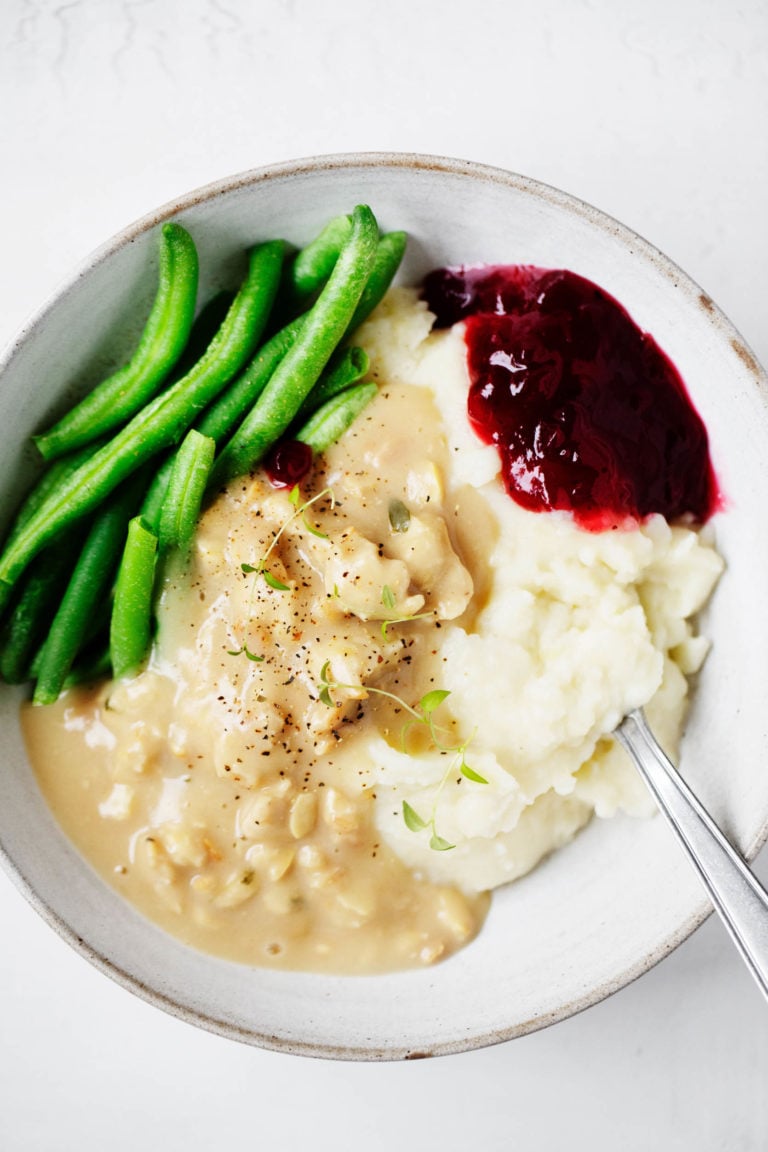 A bowl of vegan holiday food, including mashed potatoes, tempeh and gravy, green beans, and a little scoop of cranberry sauce.