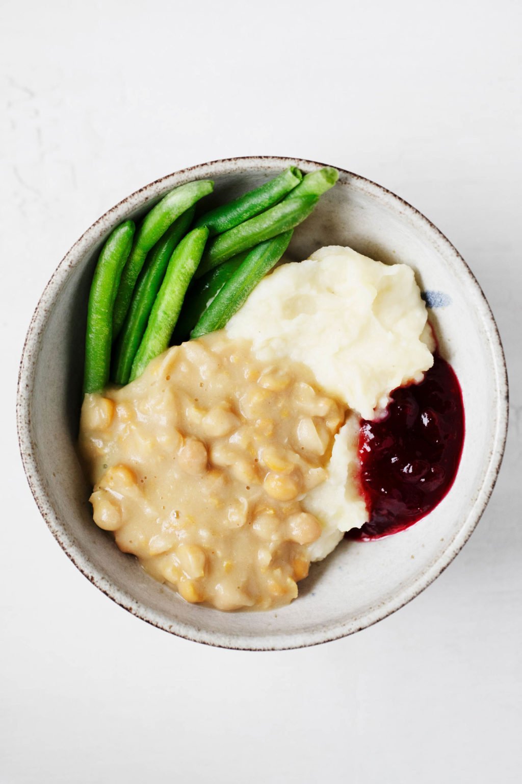 Chickpeas are paired with a savory gravy, mashed potatoes, cranberry sauce, and green beans for a filling vegan holiday bowl of food.