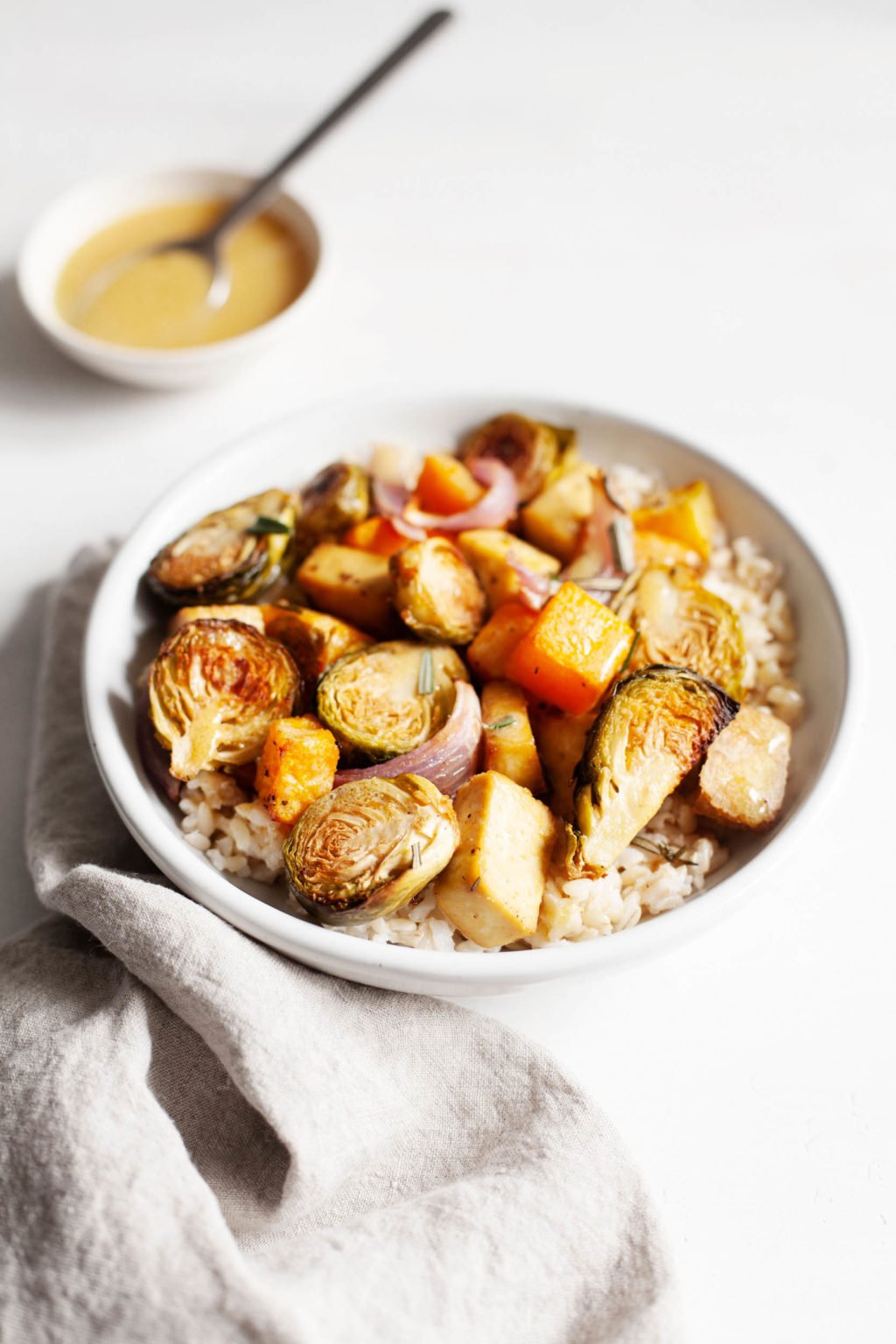 A bowl filled with Roasted brussels sprouts, butternut squash, onions, and tofu is accompanied by a small pinch bowl of vinaigrette.