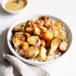 A bowl filled with Roasted brussels sprouts, butternut squash, onions, and tofu is accompanied by a small pinch bowl of vinaigrette.