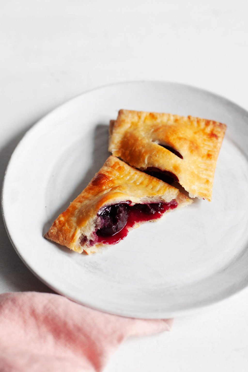 A vegan sweet cherry hand pie, freshly baked and broken apart for eating, rests on a dessert plate with a pink napkin nearby.