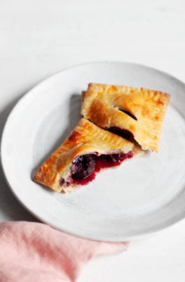 A vegan sweet cherry hand pie, freshly baked and broken apart for eating, rests on a dessert plate with a pink napkin nearby.