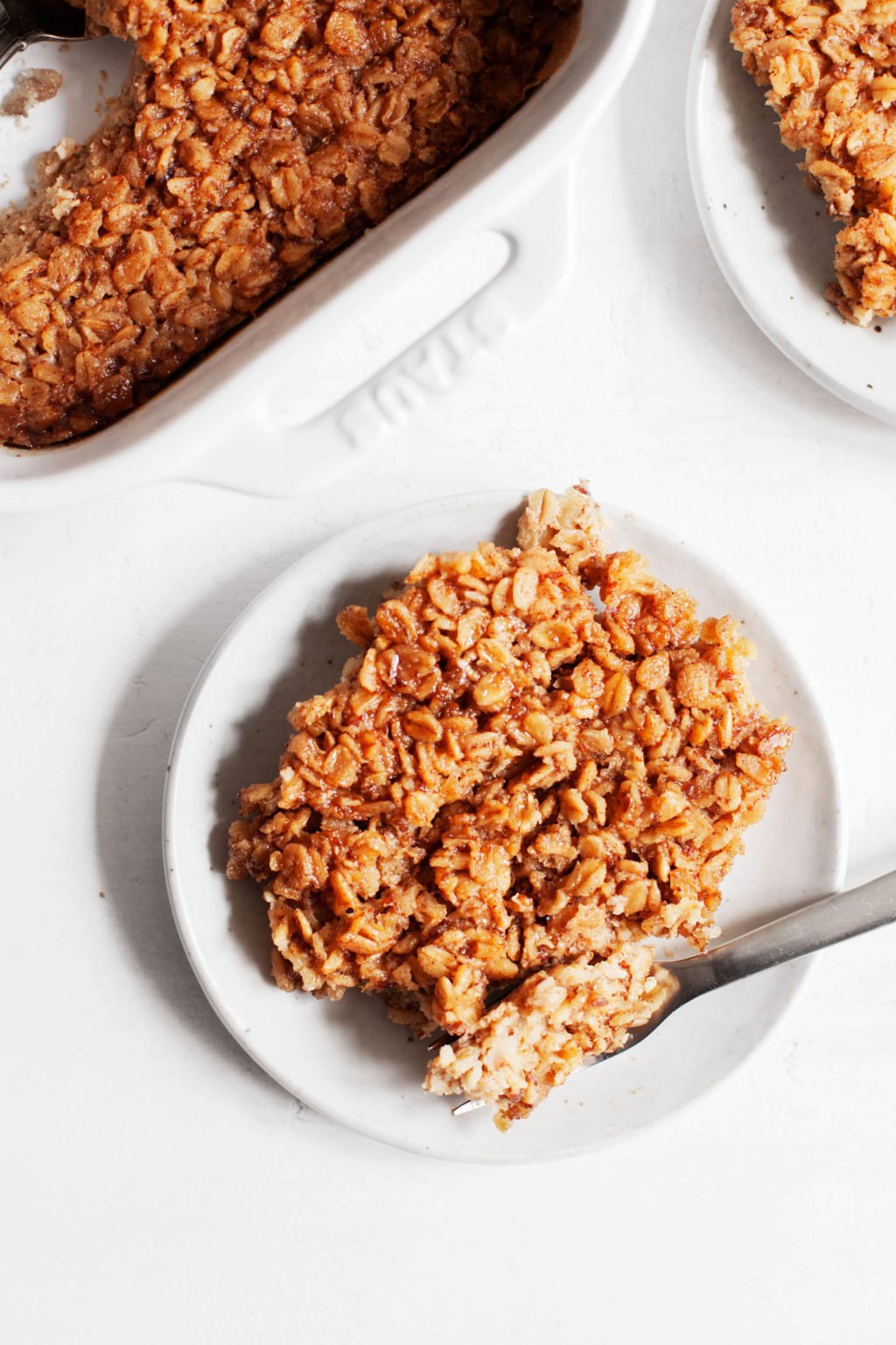 A plate of vegan, maple brown sugar baked oatmeal, with the baking dish and another serving lying nearby.