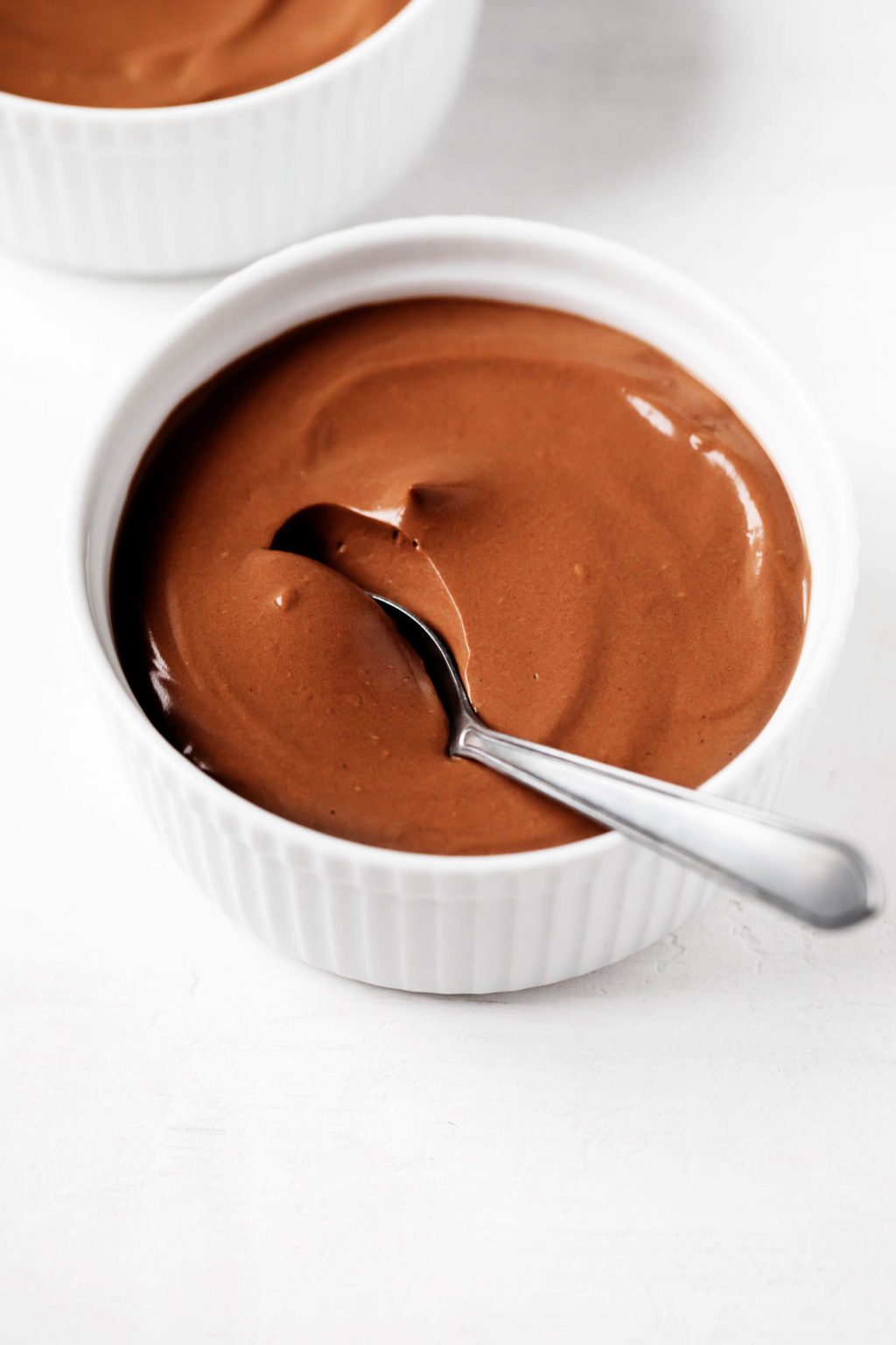 Two ramekins are filled with a creamy vegan silken tofu chocolate pudding. One has just been scooped into with a spoon. 