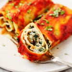 Two plant-based lasagna rolls are on a serving plate, and one of the rolls has just been sliced into.