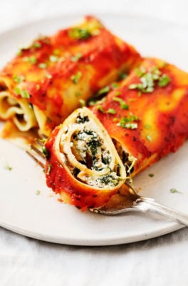 Two plant-based lasagna rolls are on a serving plate, and one of the rolls has just been sliced into.