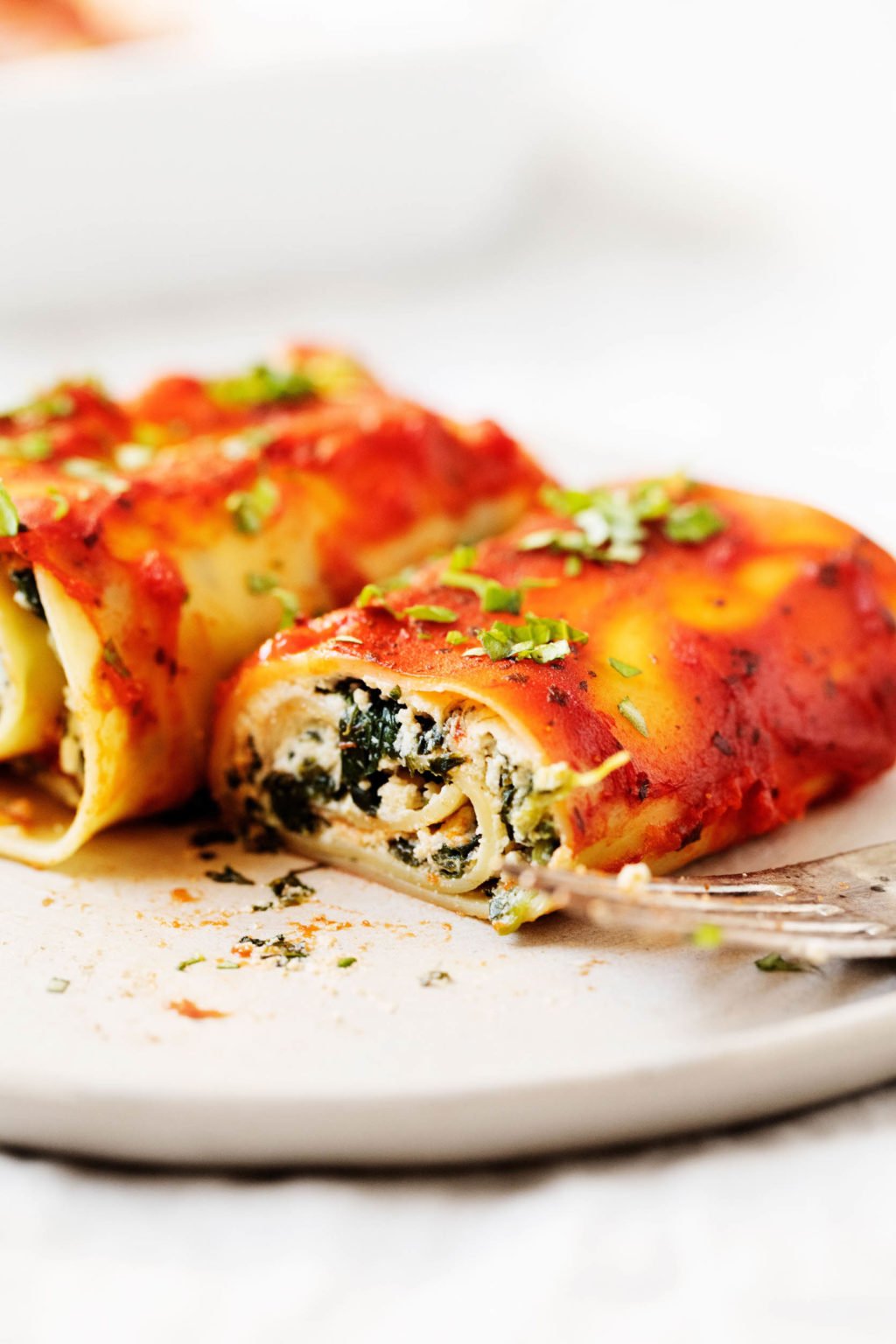 Two vegan spinach lasagna rolls sit on a dinner plate, ready to be eaten, with a fork nearby. One roll has been sliced into, revealing a creamy ricotta spinach interior.