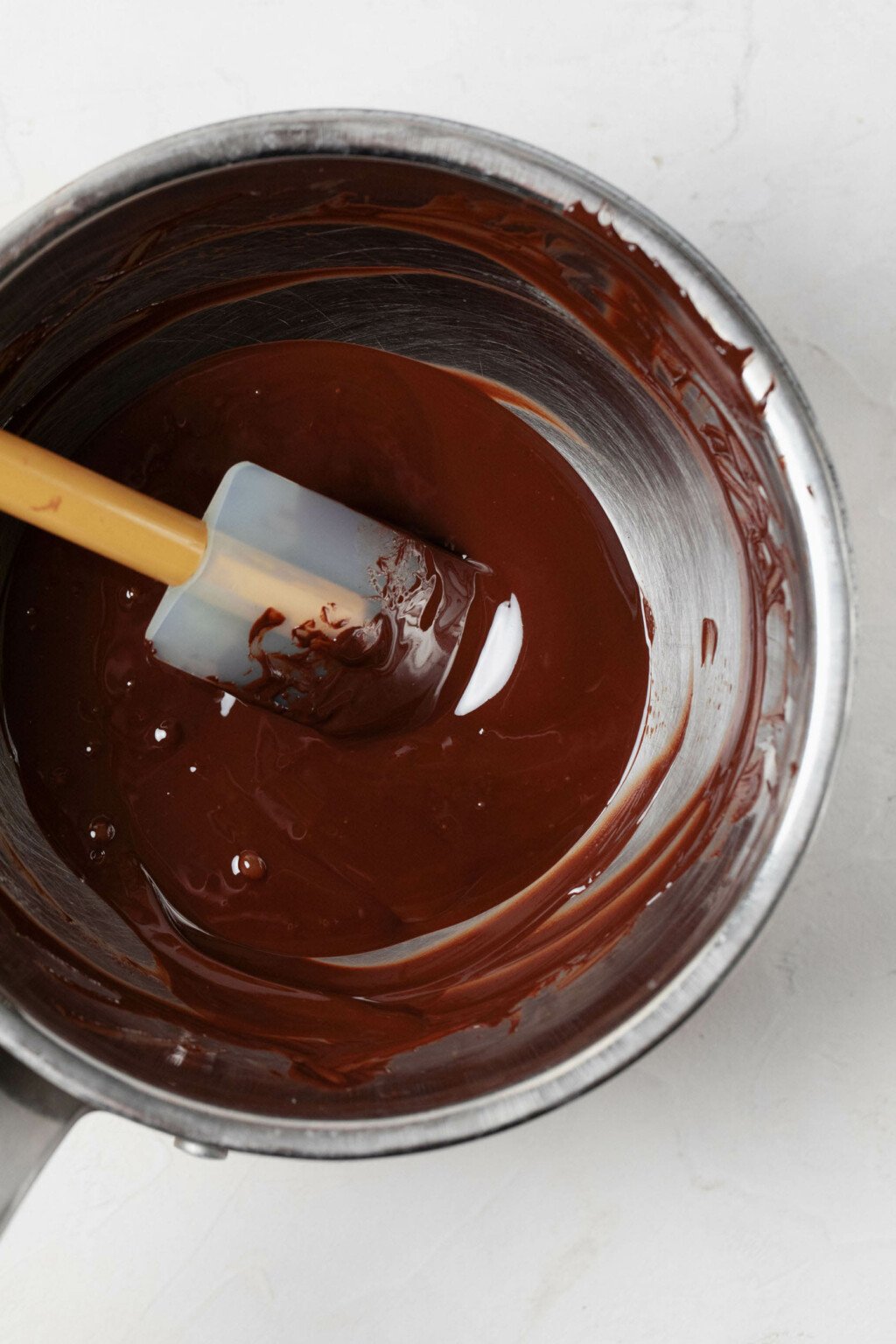 An overhead image of a metal pot, filled with shiny, melted dark chocolate. 