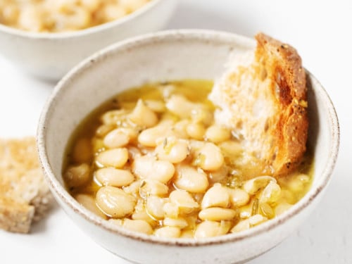 Two matching, white bowls are filled with beans, broth, and torn pieces of fresh bread.