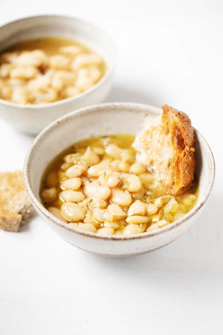 Two matching, white bowls are filled with beans, broth, and torn pieces of fresh bread.