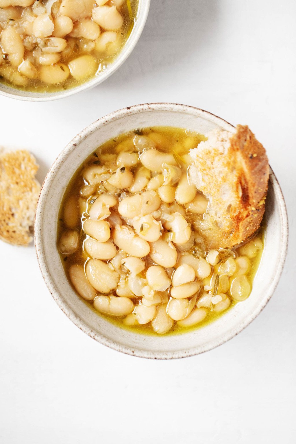 Two white ceramic bowls are filled with brothy white beans, olive oil, and small pieces of bread.