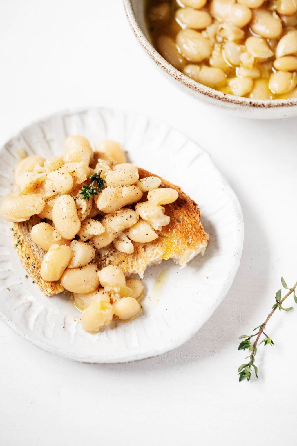 Dos pequeños platos para servir contienen tostadas cubiertas con frijoles blancos caldosos estofados. Una ramita de tomillo descansa sobre una superficie cercana.