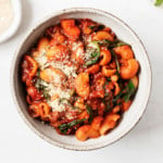 A round bowl of vegan lentil tomato pasta stew is accompanied by pinch bowls of herbs and plant-based parmesan cheese.
