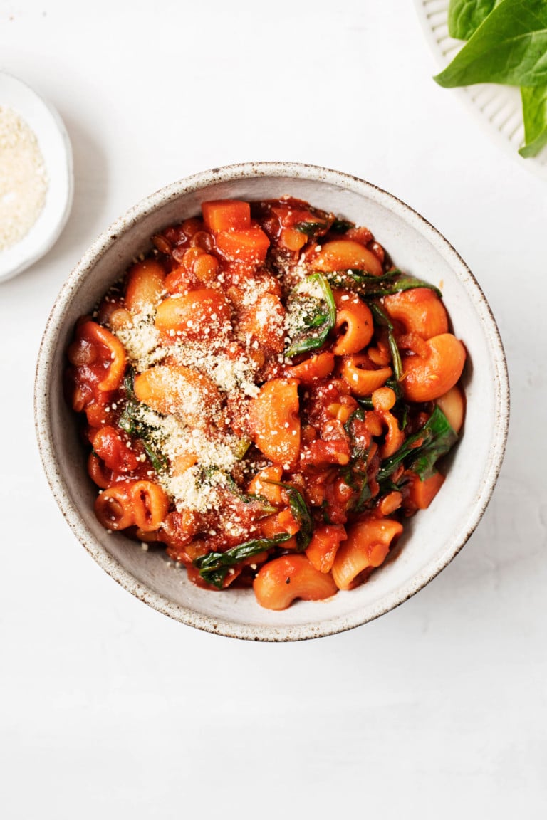 A round bowl of vegan lentil tomato pasta stew is accompanied by pinch bowls of herbs and plant-based parmesan cheese.