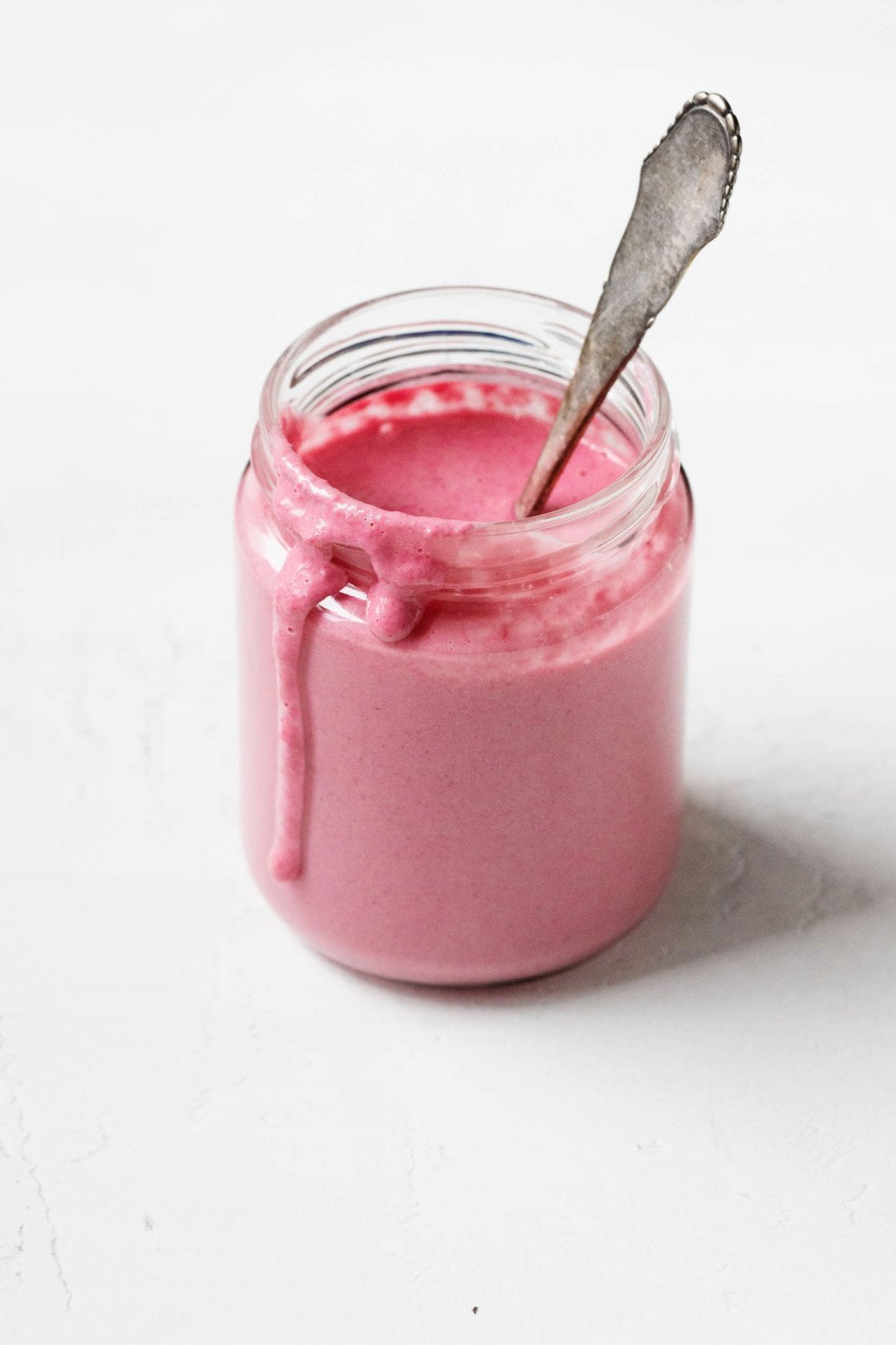 Un frasco de vidrio contiene un aderezo de remolacha tahini rosa brillante y una cuchara para mezclar.