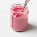 Un frasco de vidrio contiene un aderezo de remolacha tahini rosa brillante y una cuchara para mezclar.