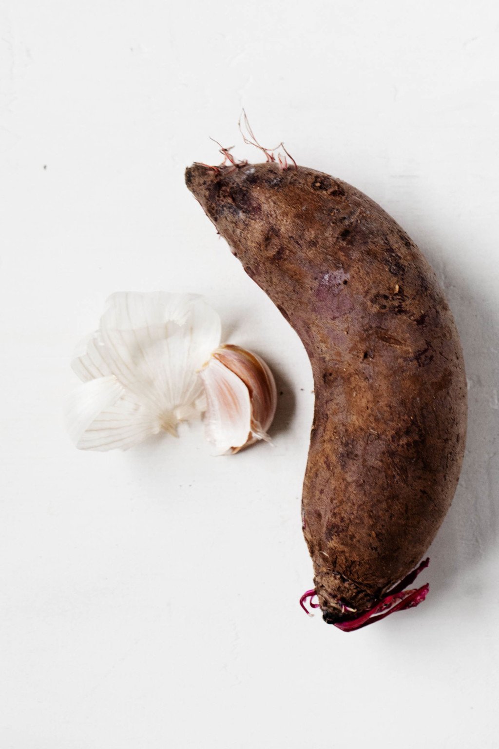 An oval shaped beet and a peeled clove of garlic on a while surface.