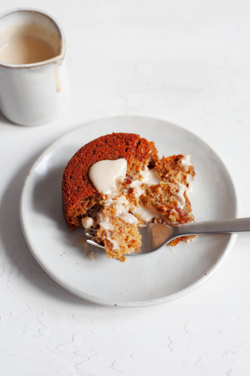 A round, vegan sticky toffee pudding has been covered in cashew cream and cut into with a serving fork. More cashew cream is in a small pitcher behind the dessert.