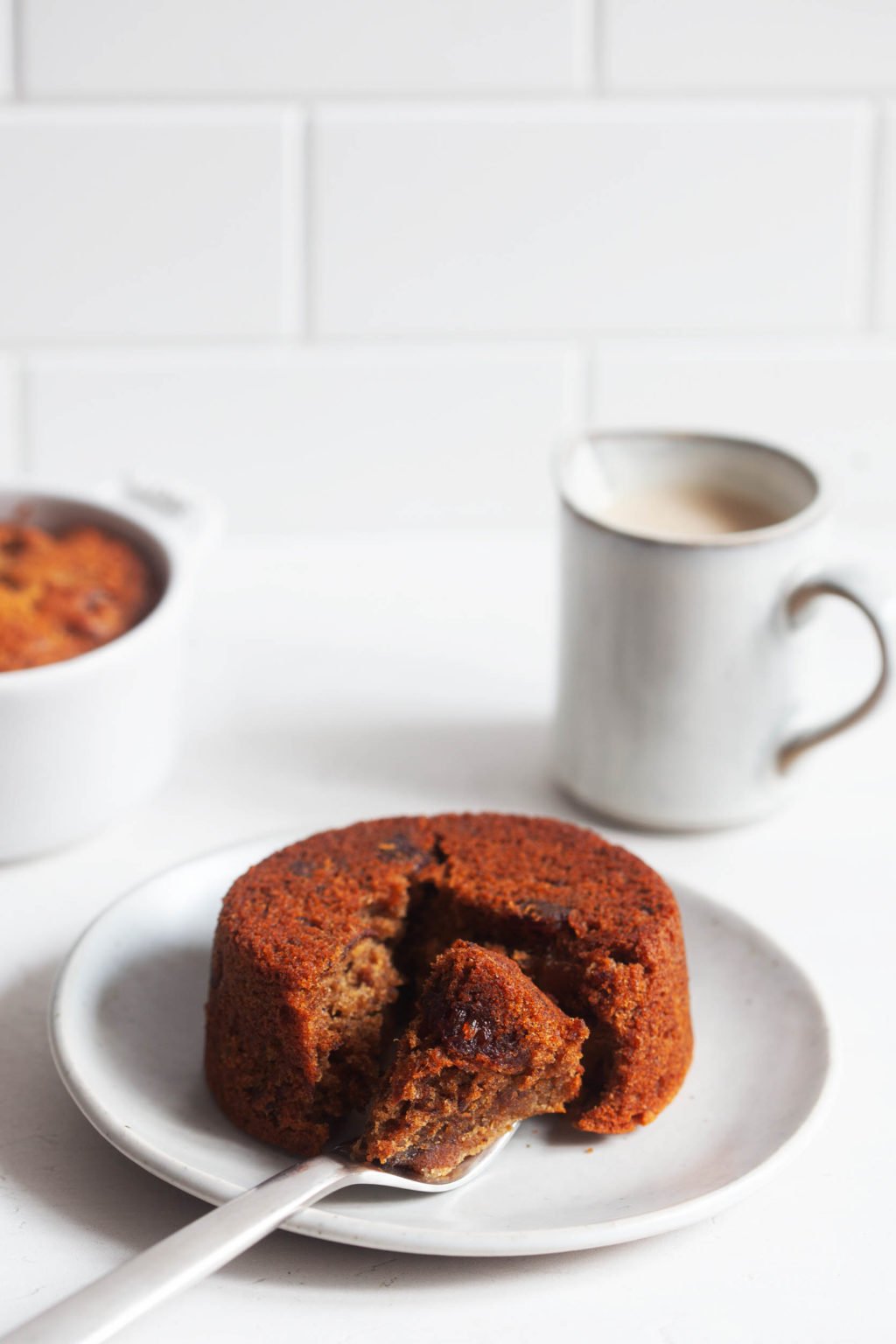 A plant-based version of sticky toffee pudding, studded with dates, sits on a dessert plate. A piece of the pudding has been cut into with a fork.