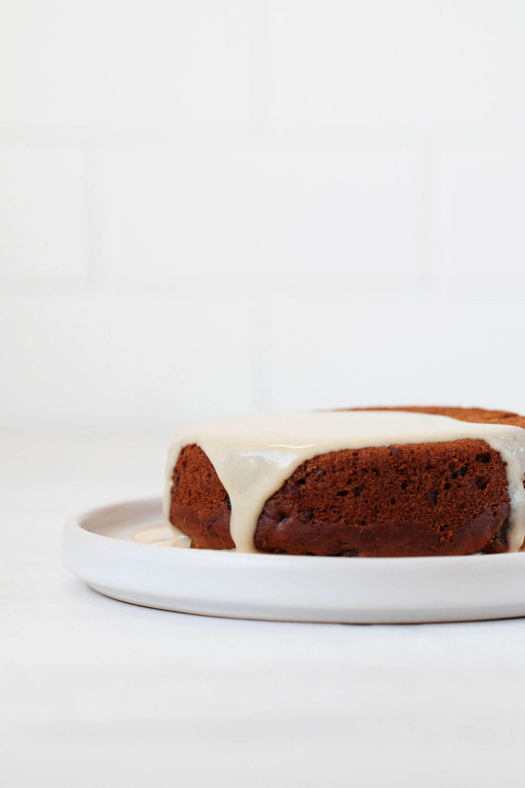 A round vegan sticky toffee pudding is resting on a small white dessert plate. It's topped with a cream sauce. There's a white brick backdrop visible behind it.