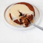 Vegan sticky toffee pudding is on a small dessert plate, with cashew cream sauce and another ramekin of pudding in the background.