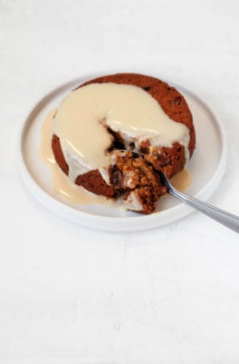 Vegan sticky toffee pudding is on a small dessert plate, with cashew cream sauce and another ramekin of pudding in the background.