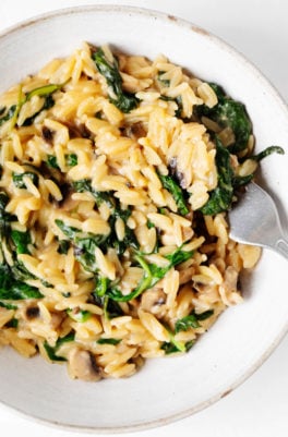 A zoomed in photograph of a bowl of creamy mushroom spinach orzo, which is accompanied by a fork.