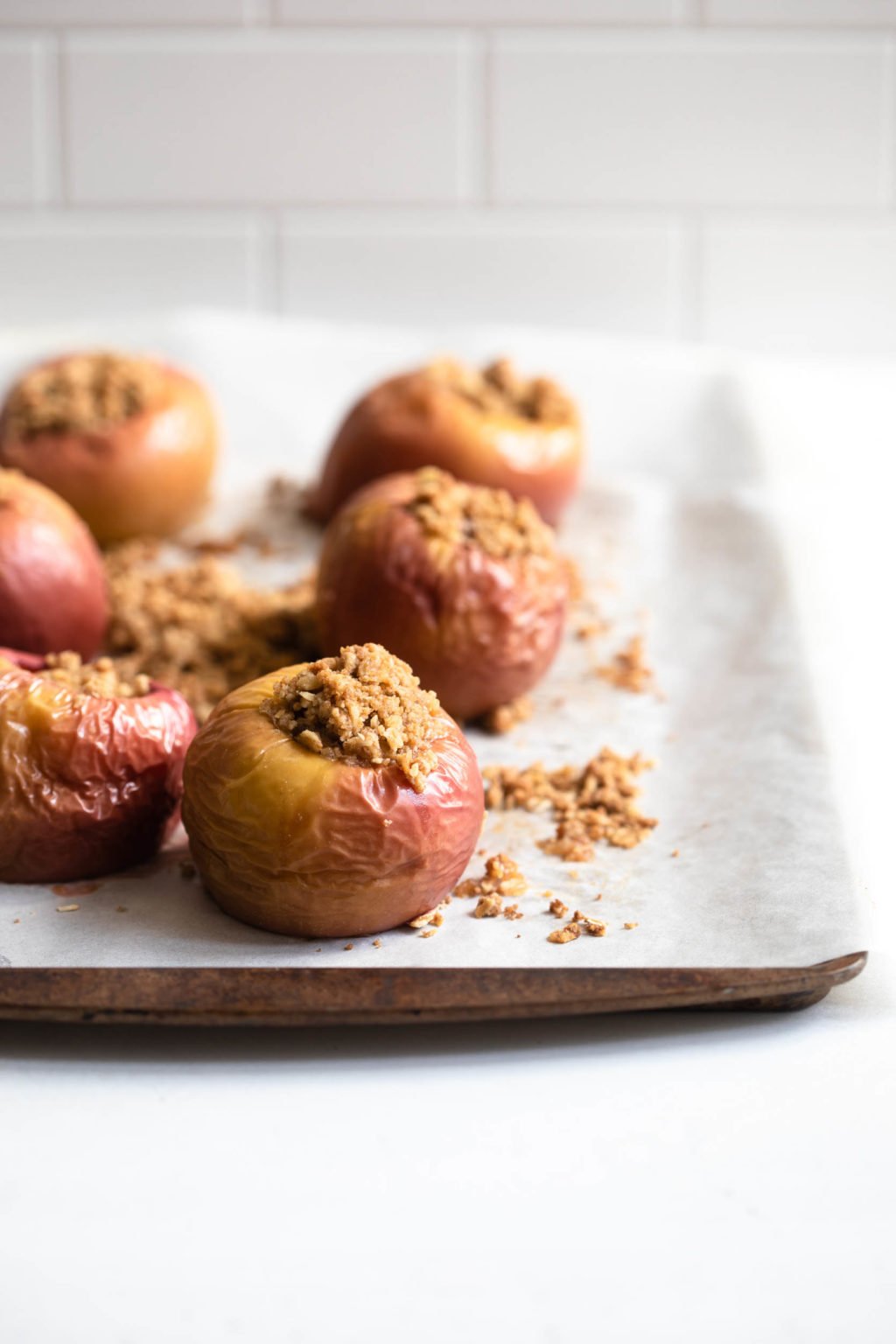 A parchment lined baking sheet holds six baked stuffed apples, with streusel crumbs on the parchment.