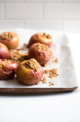 A parchment lined baking sheet holds six baked stuffed apples, with streusel crumbs on the parchment.