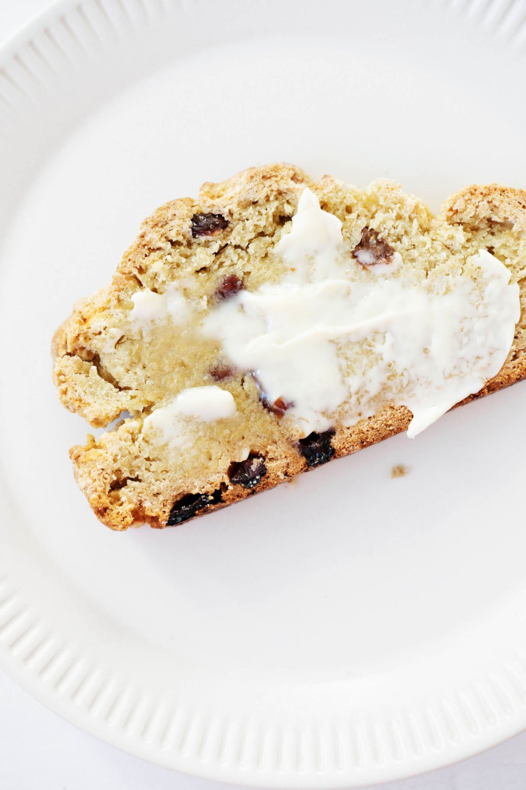 A close up photo of a slice of quickbread, which has been topped with a schmear of butter. It rests on a fluted white ceramic plate.