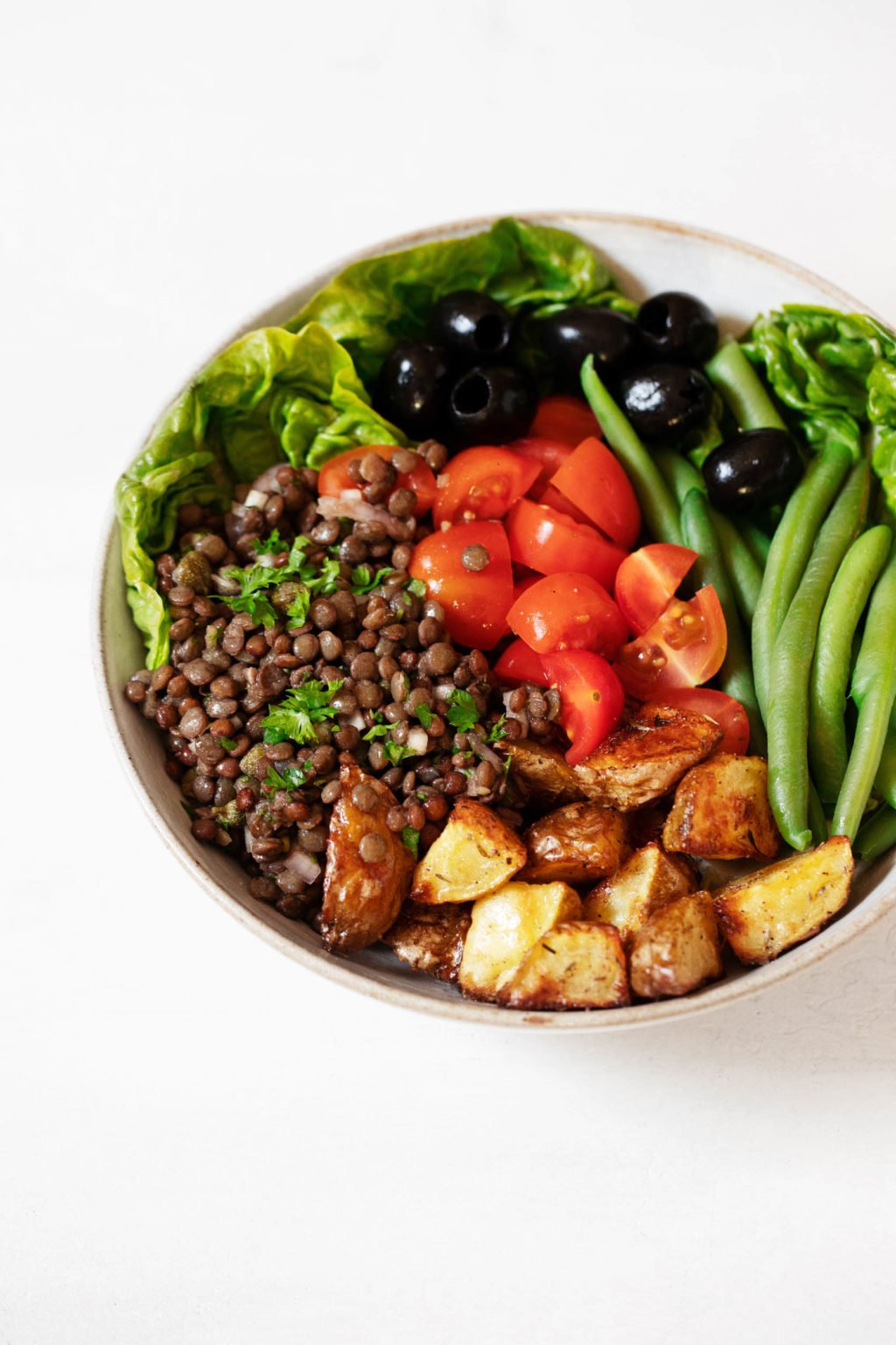 A white bowl has been piled with cooked French lentils, roasted potatoes, tomatoes, olives, and green beans.