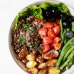 A round, white salad bowl has been filled with the colorful ingredients for a vegan Niçoise salad, including tomatoes, lentils, potatoes, and green beans.