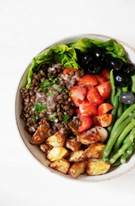 A round, white salad bowl has been filled with the colorful ingredients for a vegan Niçoise salad, including tomatoes, lentils, potatoes, and green beans.