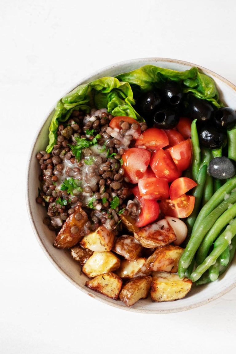 A round, white salad bowl has been filled with the colorful ingredients for a vegan Niçoise salad, including tomatoes, lentils, potatoes, and green beans.