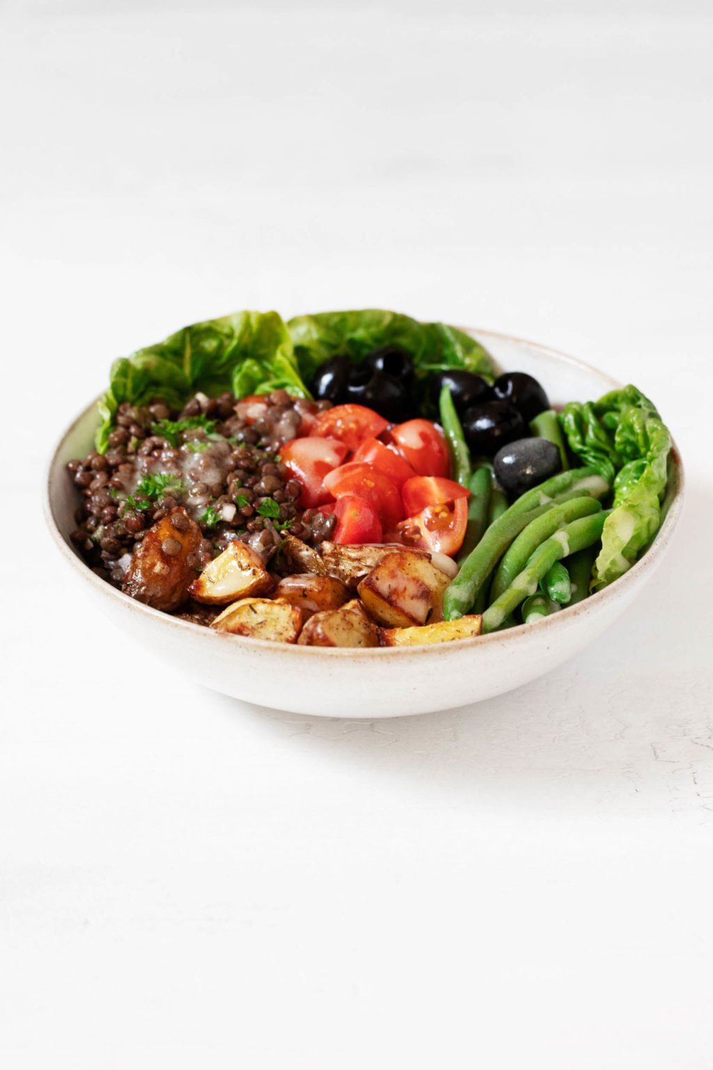 An angled photograph of a round bowl of vegan lentil Niçoise salad, which is resting on a clean white surface.