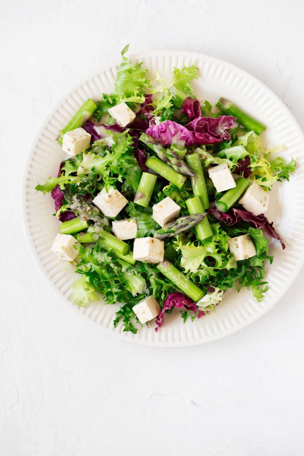 Se sirve una ensalada de primavera brillante y vibrante en un plato de cerámica blanca estriada. Una mezcla de tofu marinado es parte de la ensalada. 