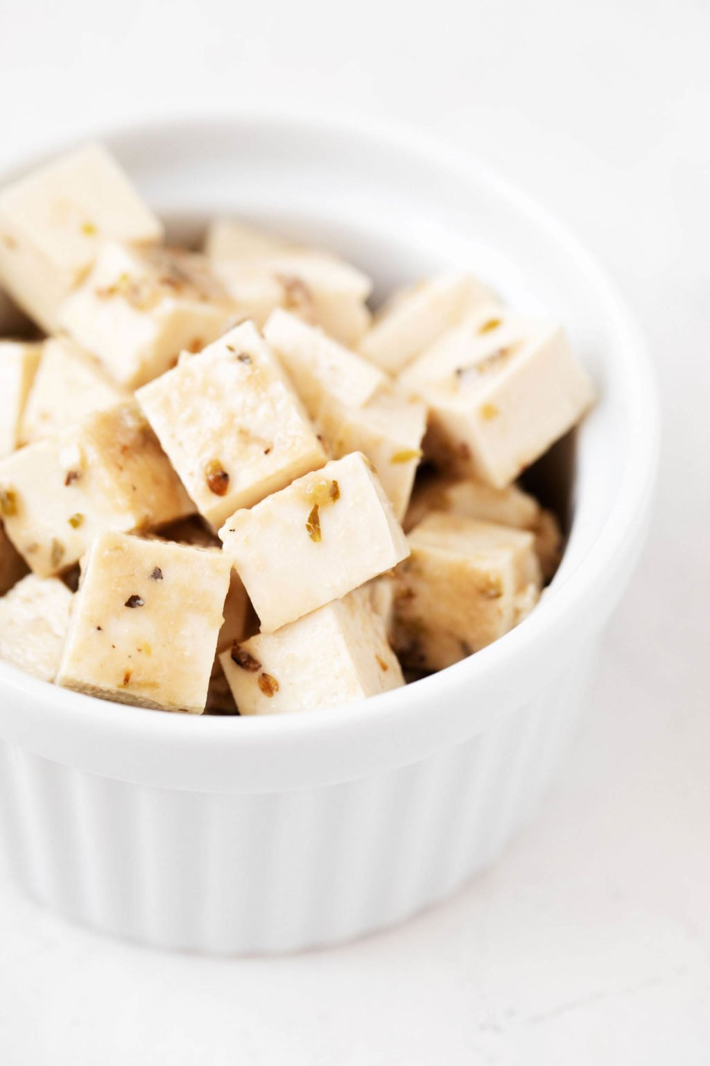 A round, white ramekin has been filled with small cubes of a vegan herbed tofu feta cheese.