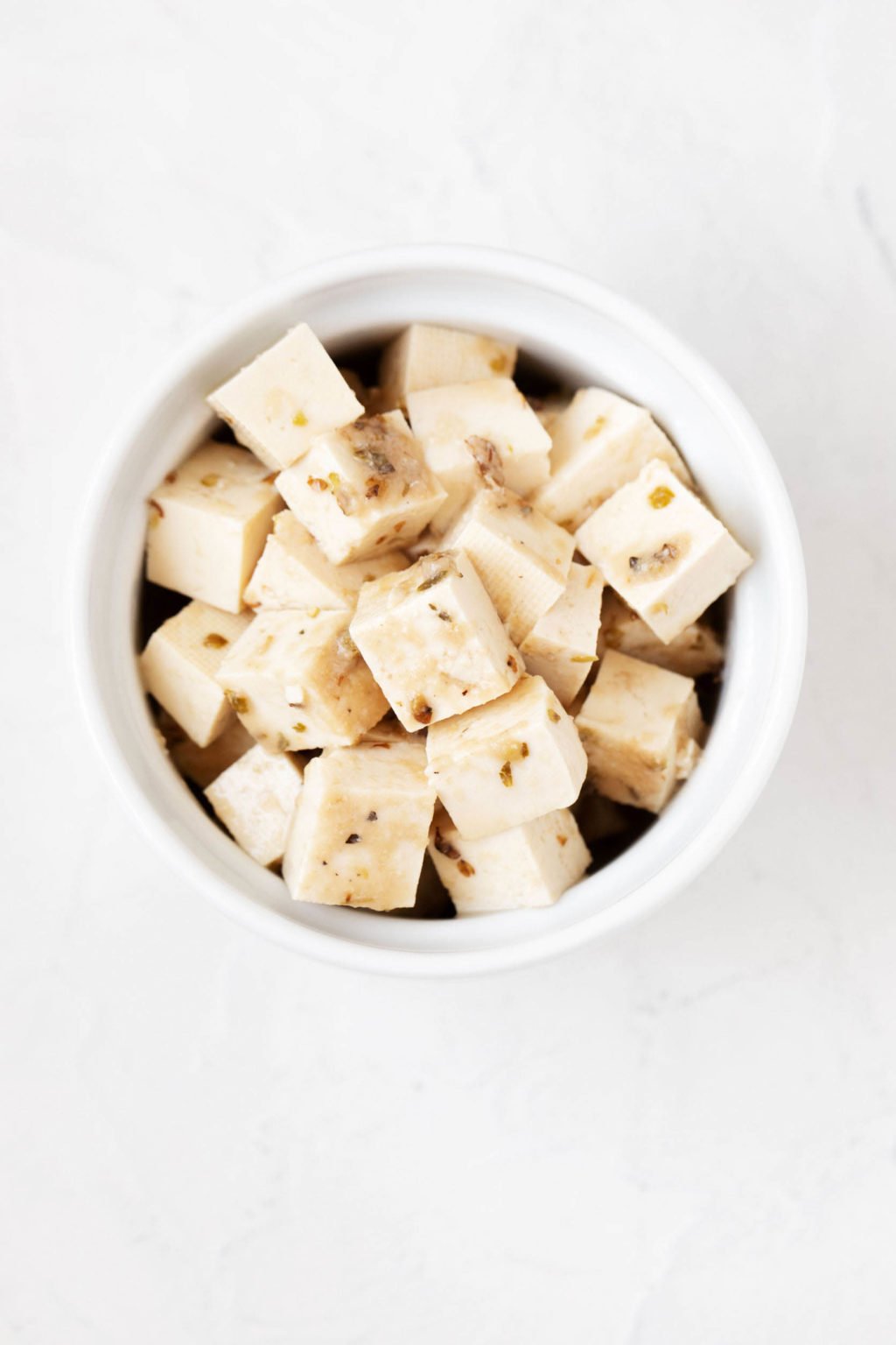A small, white round ramekin has been filled with cubes of marinated tofu and dried herbs. It sits on a white backdrop.