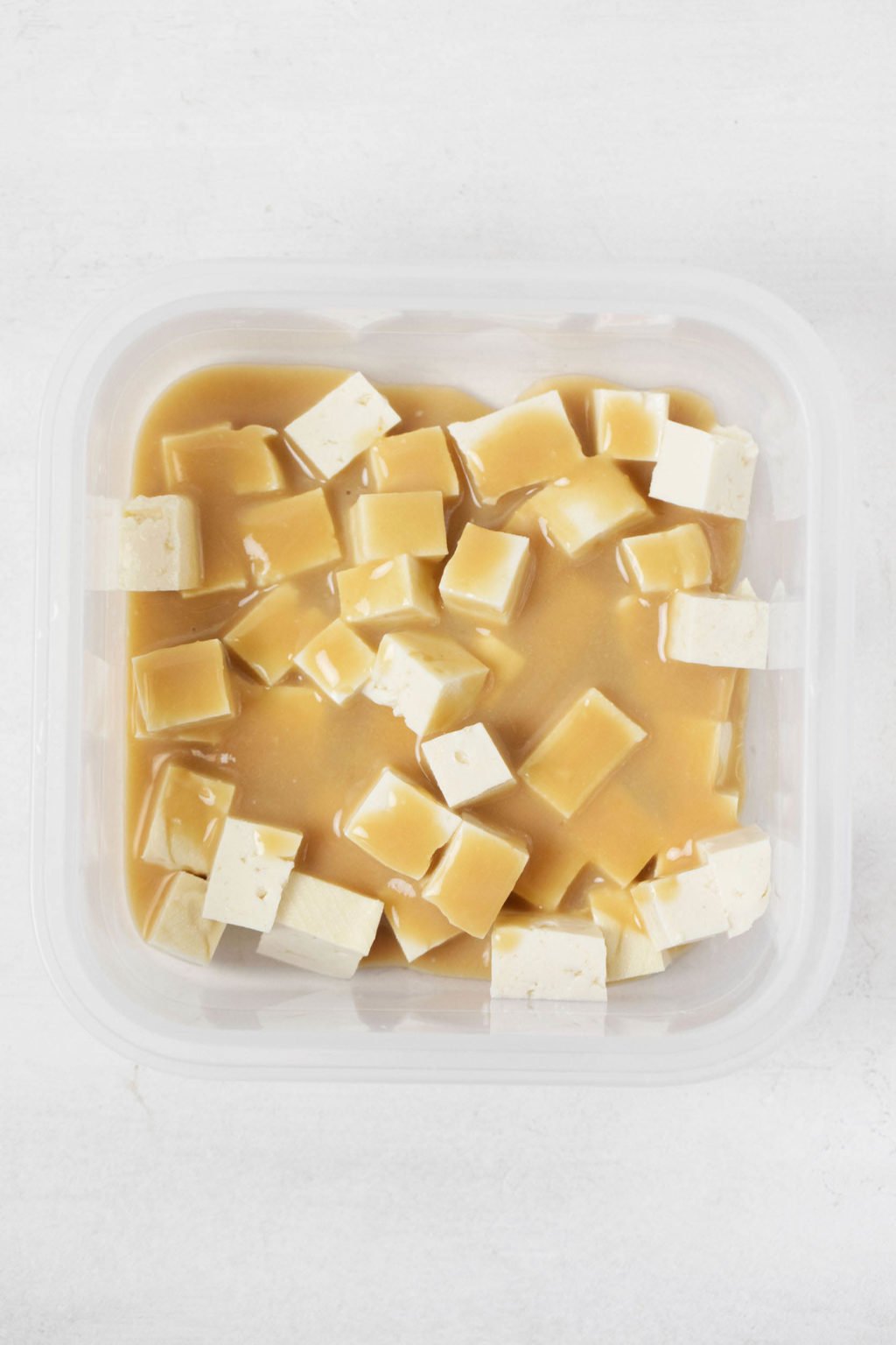 Cubes of tofu are being submerged in a thick marinade. They're held in a clear storage container.