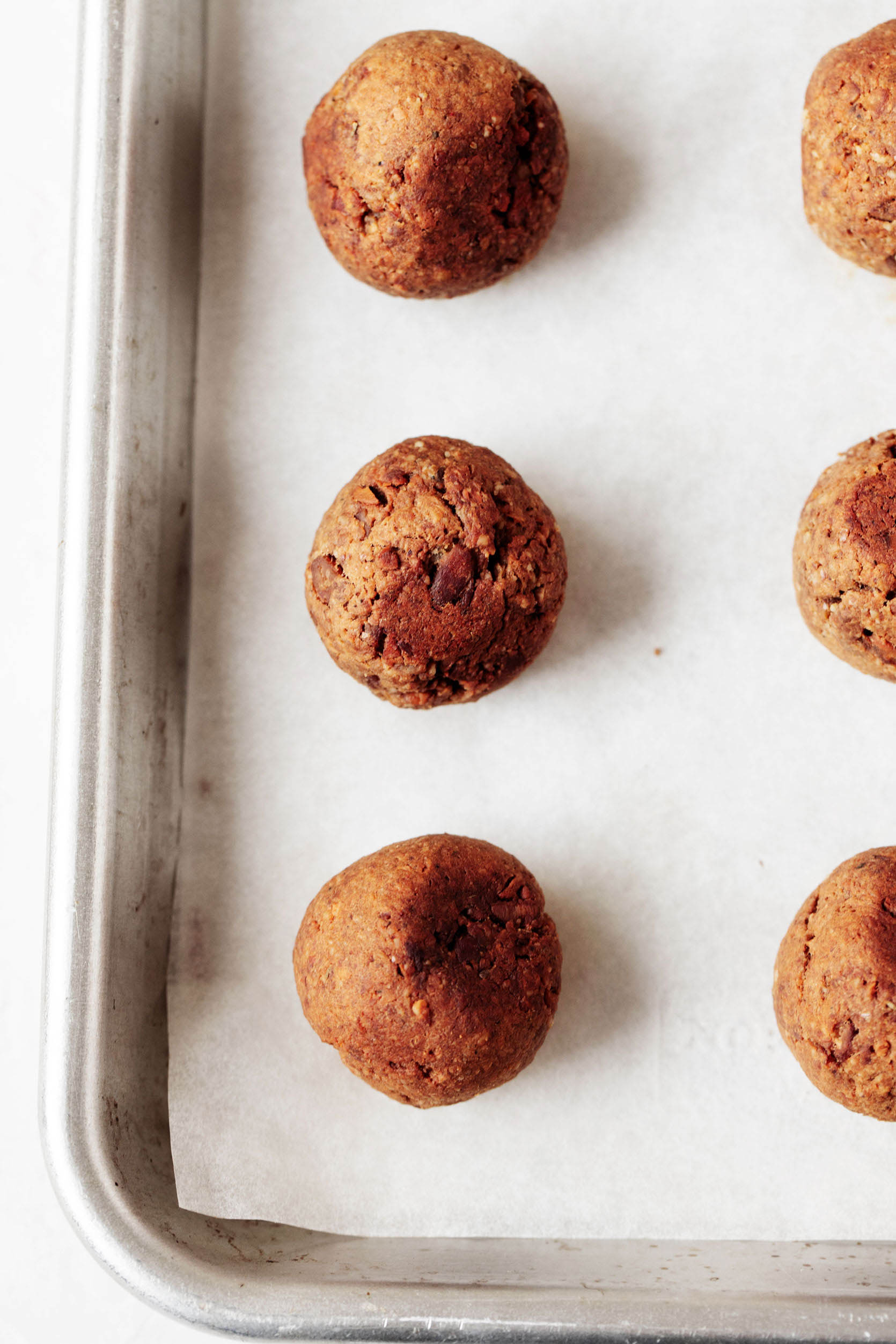 A silver rimmed baking sheet is covered with a sheet of parchment and freshly baked tempeh meatballs.