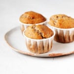 Three vegan banana walnut muffins are arranged on a white ceramic plate. The plate rests on a white surface.