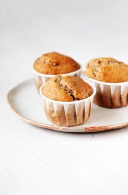Three vegan banana walnut muffins are arranged on a white ceramic plate. The plate rests on a white surface.