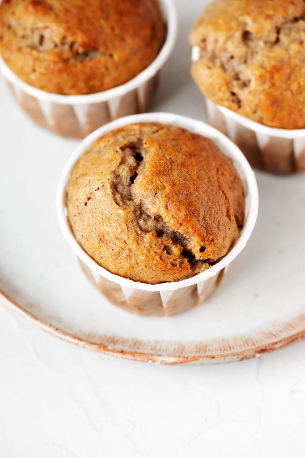 An angled, overhead image of vegan banana walnut muffins. They're lined with white paper liners and resting on a white dessert plate with a tan rim.