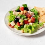 A white fluted plate holds a vegan Greek salad with chickpeas and pita wedges. It rests on a white surface.