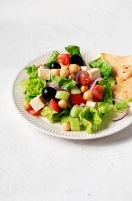 A white fluted plate holds a vegan Greek salad with chickpeas and pita wedges. It rests on a white surface.