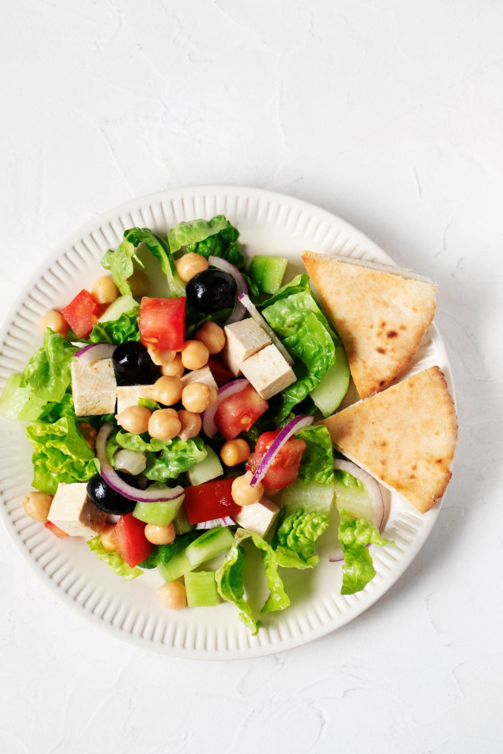 An overhead image of a crisp green salad with romaine lettuce, tomatoes, olives, red onions, and pita wedges. It's served on a white, rimmed plate.