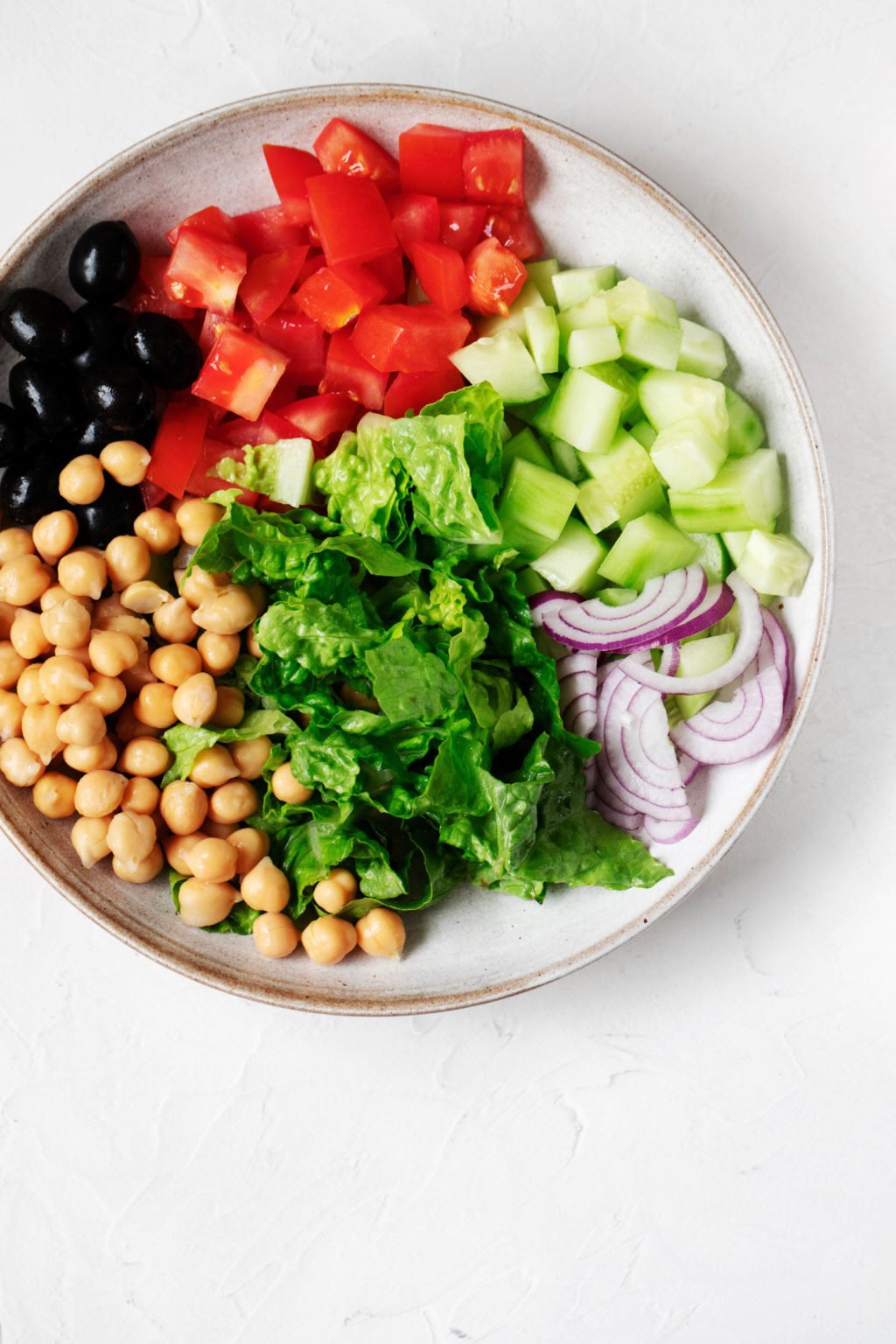 A large, white serving bowl is filled with the ingredients for a chickpea salad. It rests on a white surface.