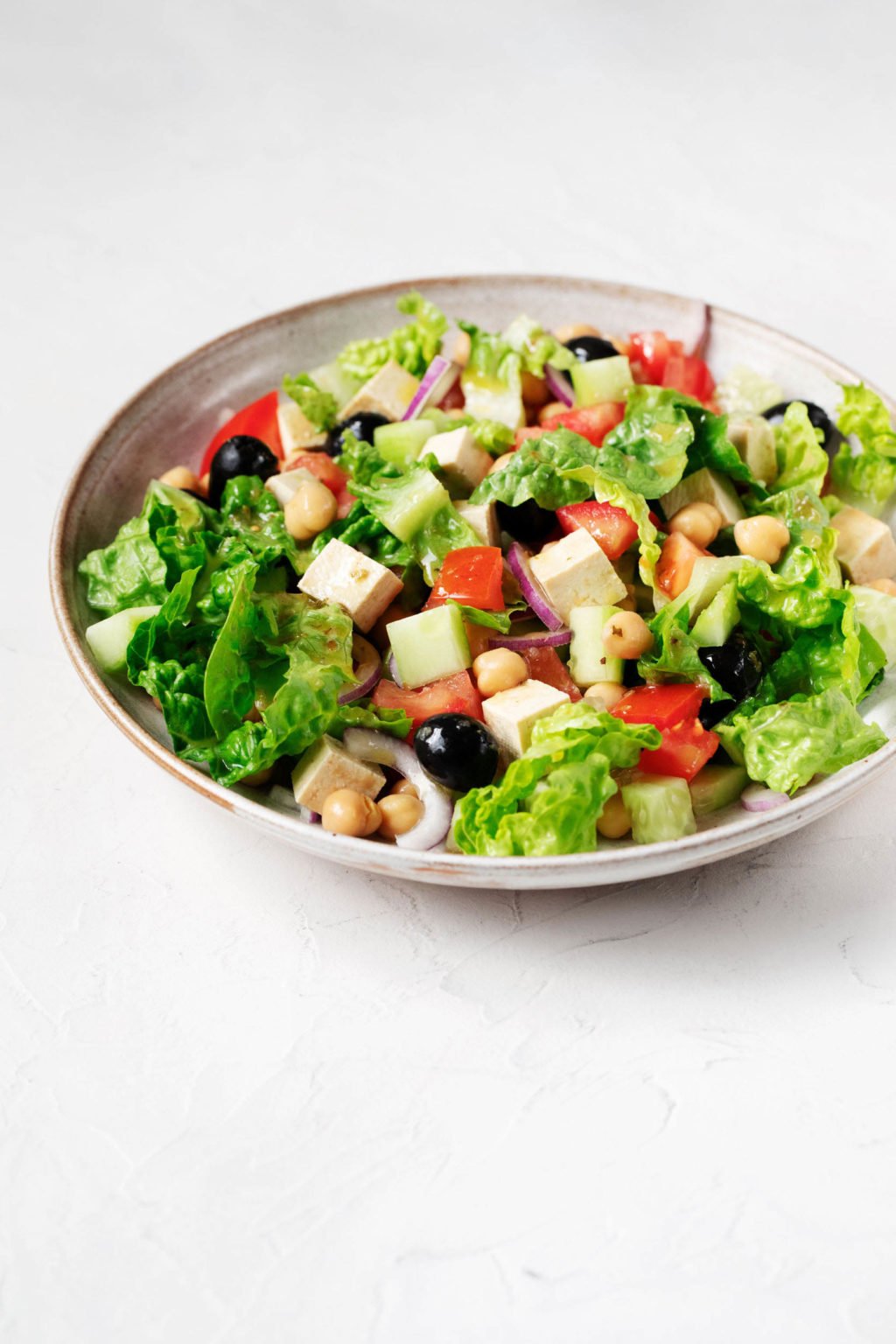 A vegan Greek salad, prepared with tofu feta and olives, is served in a large, round bowl. Crisp green romaine lettuce is folded into the salad.