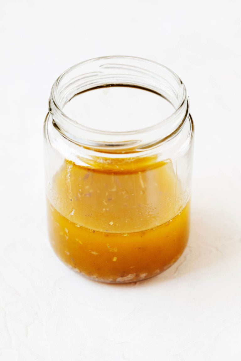 A wide-mouthed, glass jar is holding a vinaigrette salad dressing. It rests on a white surface.
