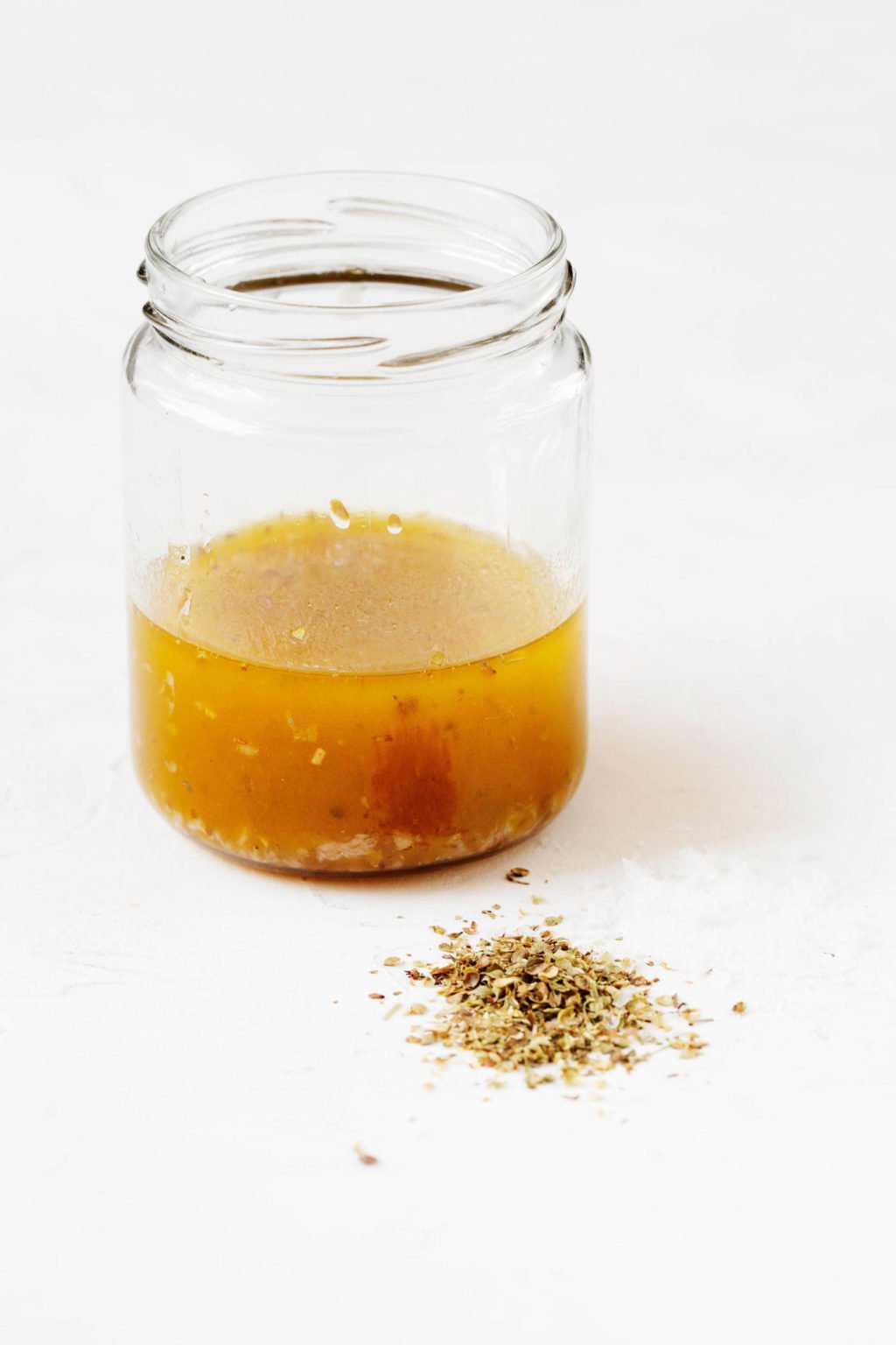 An angled photograph of a Greek vinaigrette dressing in a mason jar, with a pat of dried oregano on a white surface in front of it.